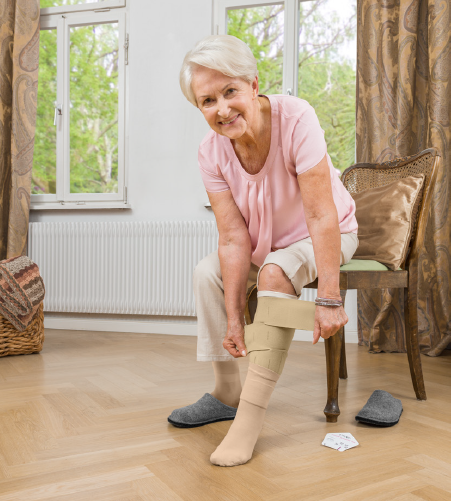 Elderly woman seated on chair applying circaid juxtalite adjustable compression wrap to her lower leg at home